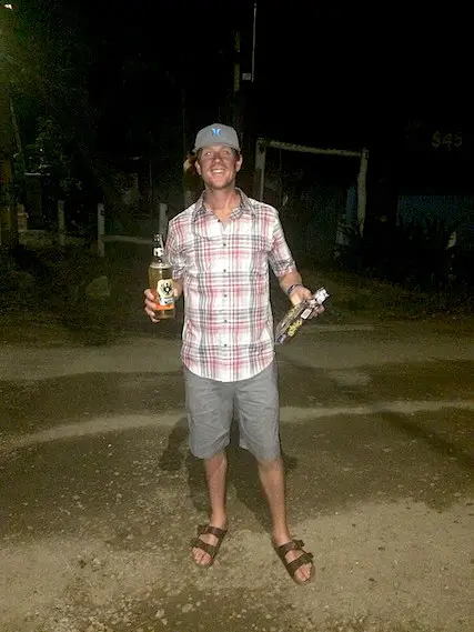 Man walking down a dirt street after dark with a large beer and bag of cookies.