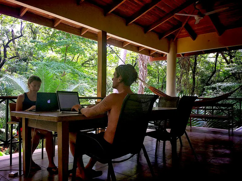 Man and woman working on laptops on a large verandah surrounded by palm trees and jungle in Nosara, Costa Rica.