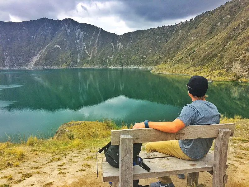 Man sitting on a bench looking at the emerald crater lake, Laguna Quilotoa, surrounded by mountains in Ecuador.