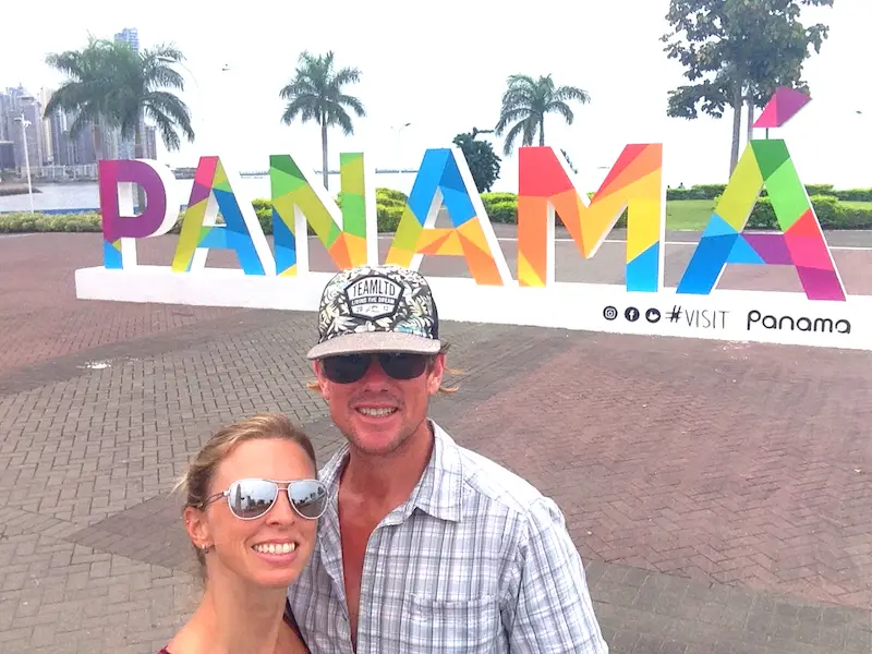 Couple standing in front of colourful Panama sign on Cinta Coster with palm trees in distance, Panama City.