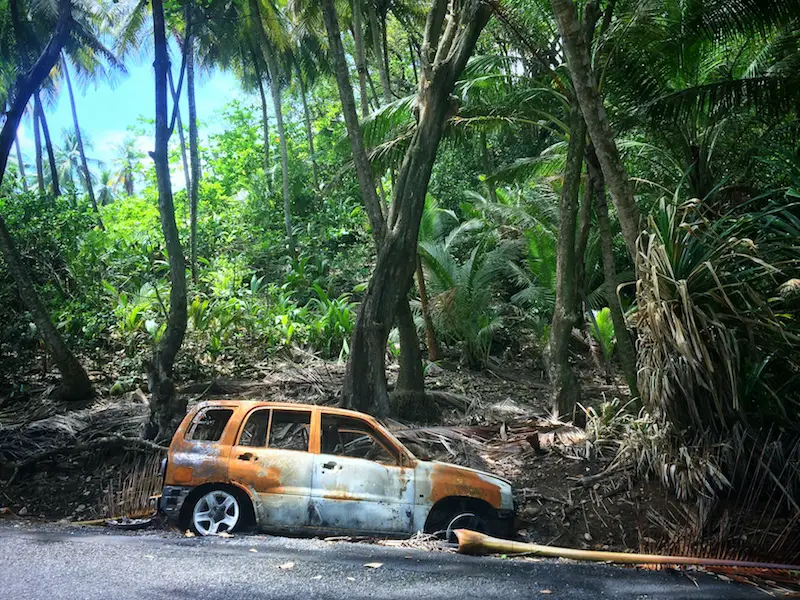 Rusted shell of a car on the side of the road surrounded by jungle in Dominica.