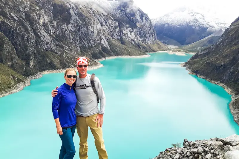 A couple standing in front of a bright turquoise lake between snow-capped mountains near Huaraz, Peru.