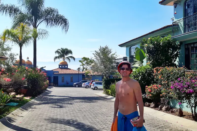 Man walking among palm trees and colourful houses with a book and towel wearing a swimsuit.
