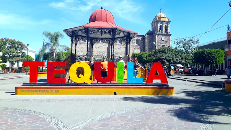 Four people standing behind the bright large letters that spell "Tequila" in Mexico.