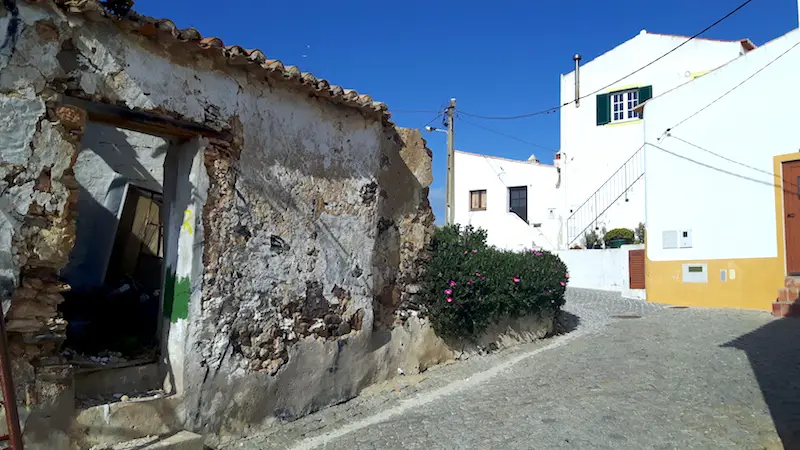 Old building beside a whitewashed one on a winding street in Carrapateira, Portugal.