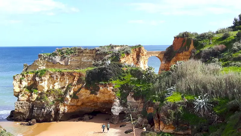 A beach surrounded by high orange cliffs with a tunnel that two people are entering in Lagos, Portugal.
