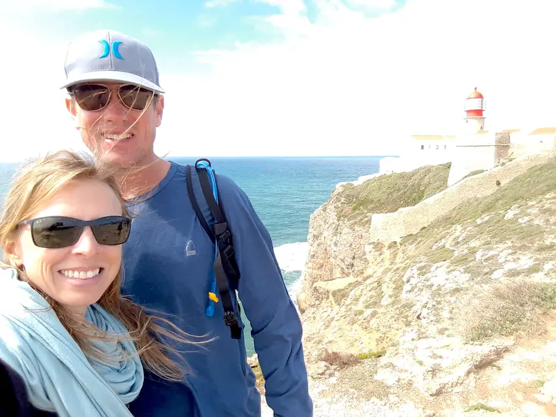 Man and woman with cliffs topped with a lighthouse behind in Portugal.