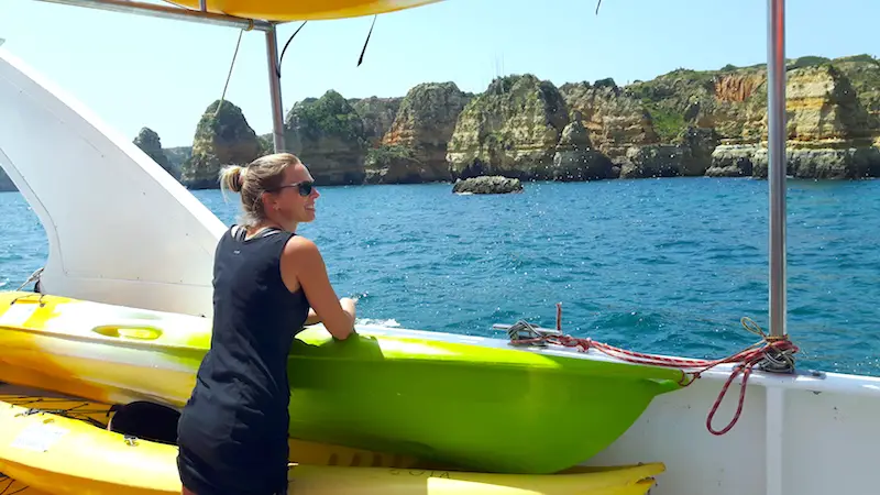 Woman leaning on stack of kayaks on the back of a boat looking at Lagos cliffs, Portugal.