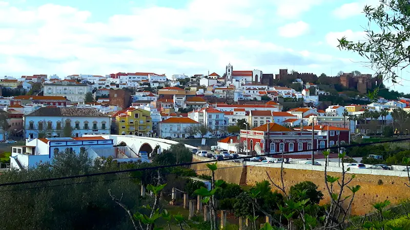 Small town of white buildings with red tiled roofs climbing a hillside across a river in Portugal.