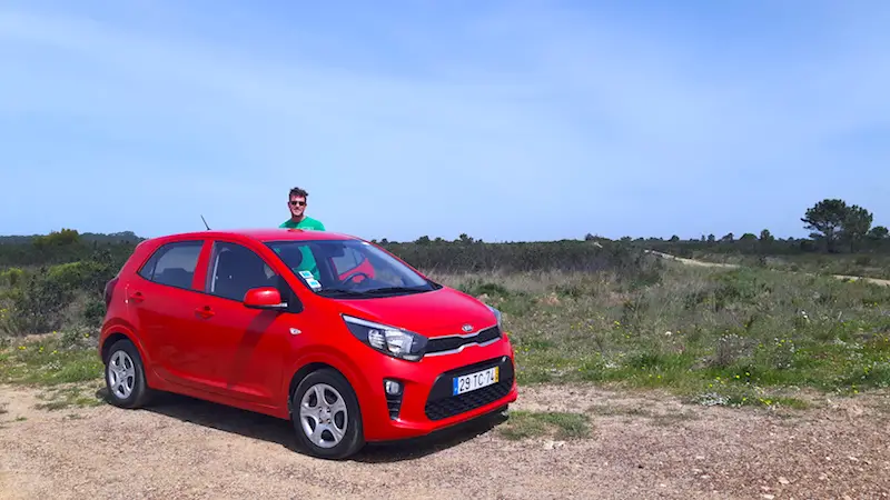 Man standing beside a bright red hatchback rental car in a field in Portugal.