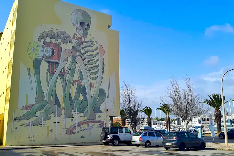 Side of multi-storey building covered in giant skeleton street art in Lagos, Portugal.