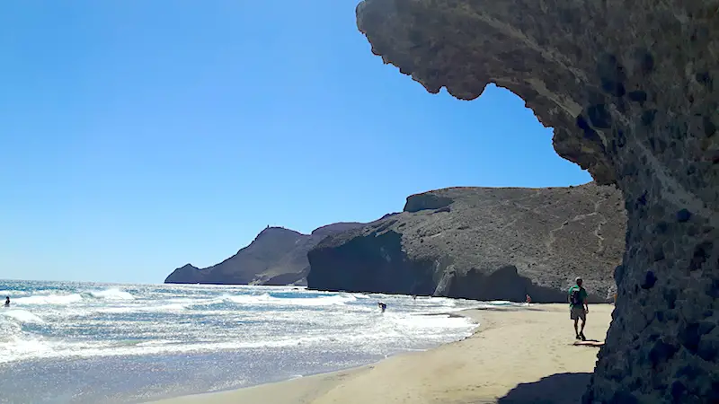 Man walking along a Mediterranean beach with a volcanic rock shaped like a wave towering over him at Playa Monsul in Cabo de Gata, Spain.