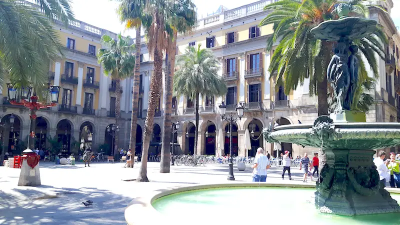 Central fountain surrounded by people, palm trees and traditional architecture in Plaza Real in Barcelona, Spain.