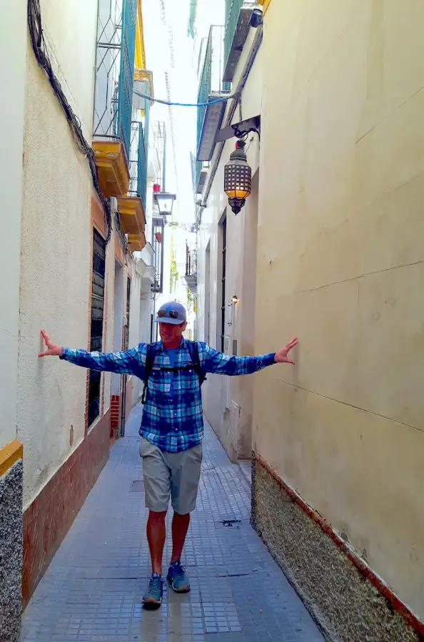 Man touching buildings on each side of a very narrow road in Seville Spain.