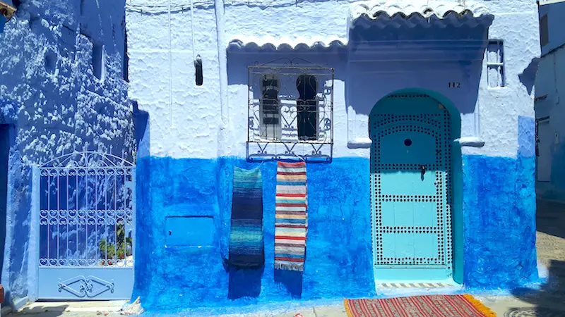 turquoise door with colourful blankets hanging under a window in Chefchaouen, Morocco's Blue City.