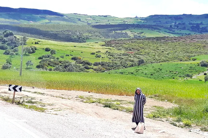 Man in traditional dress waiting on the side of the highway with mountain views behind in Morocco.