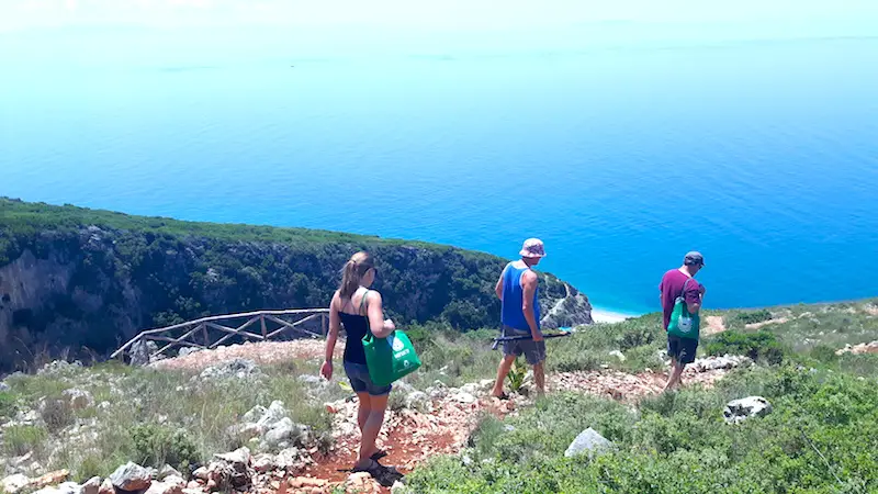 Three people hiking down a rocky trail beside a deep gorge with the bright blue Mediterranean in front in Albania.