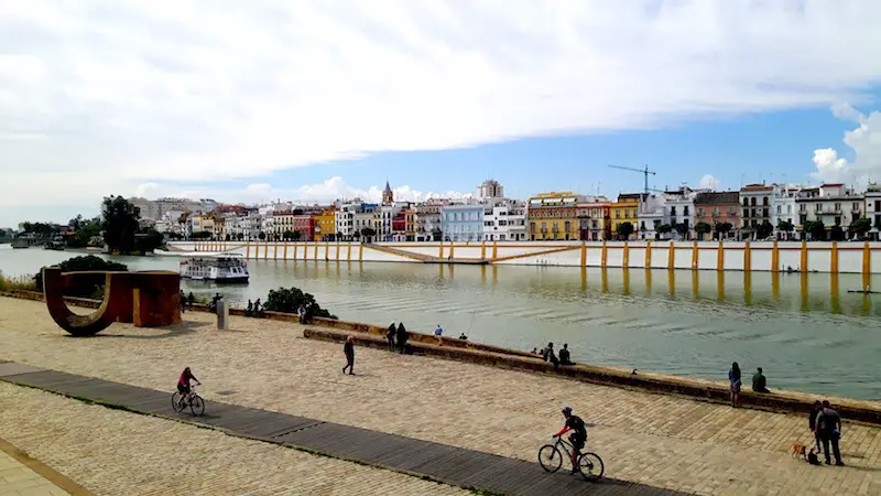 Path running along a river with people cycling and colourful buildings on the other side in Seville, Spain.