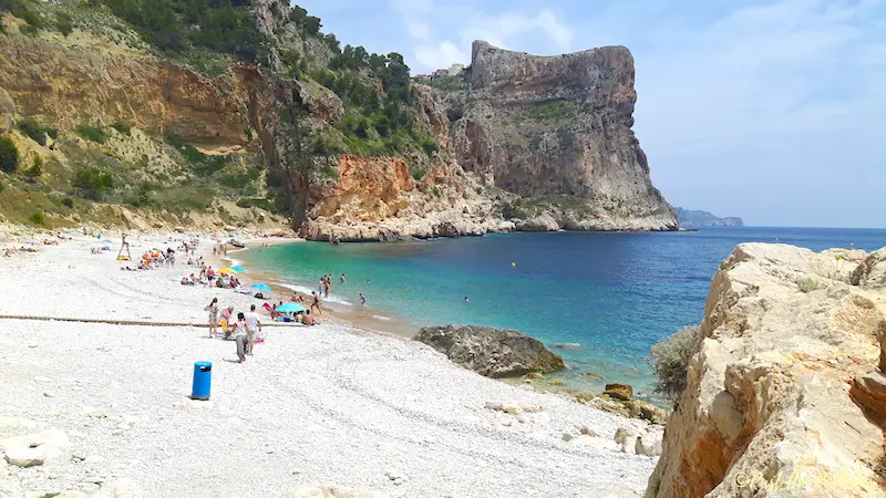 Turquoise water with orange cliffs and pebble beach at Playa Benitachell in Spain.