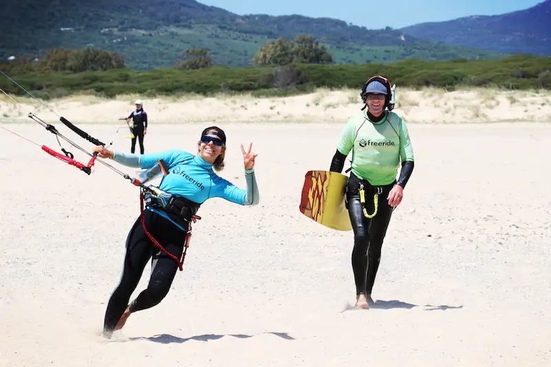 Kitesurfing instructor with blue Freeride Tarifa rash guard and a student with a green one carrying a kite board along the beach in Spain.