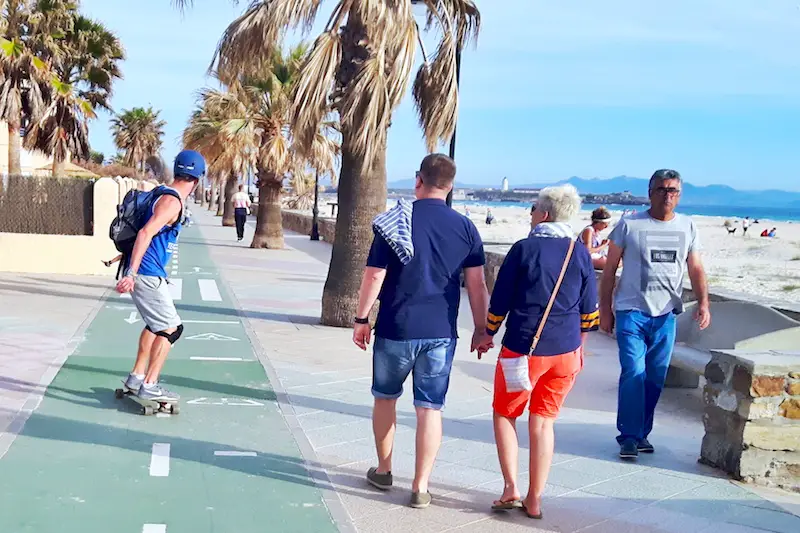 Man long boarding with helmet on and other people walking along the boardwalk in Tarifa, Spain.