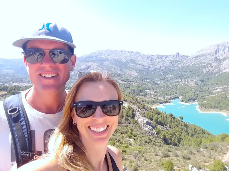 Couple smiling with bright blue reservoir behind surrounded by mountains in Castell de Guadalest, Spain.