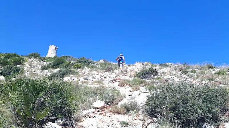 Man hiking up a steep, rocky hillside to an old watchtower at the summit in Denia, Spain.