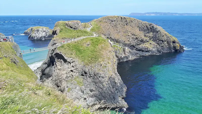 A rope bridge connecting a small rock island from mainland Northern Ireland across bright turquoise water.