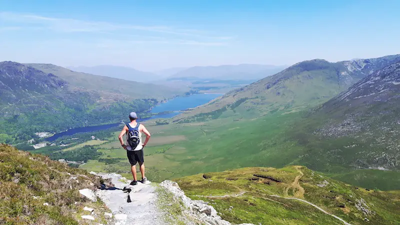 Man looking out across rolling green hills and a blue lake from the top of Diamond Hill hike, Connemara National Park, Ireland.