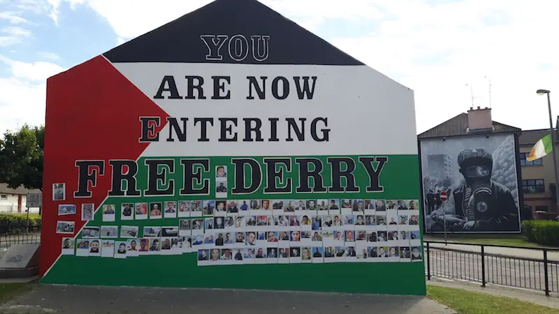 Wall painted with text: You are now entering Free Derry and a mural on a building behind from the 'troubles' in Northern Ireland.