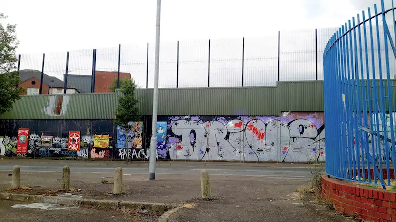 Graffiti covered concrete wall with tall wire fence above. The peace wall in Belfast, Northern Ireland.