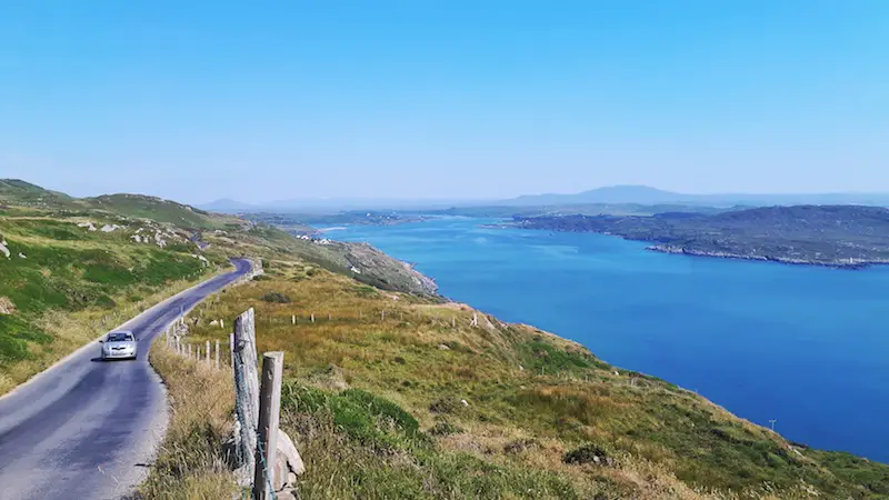 An oncoming car along the narrow winding road running on the edge of a hill down to the bright blue water in Ireland.