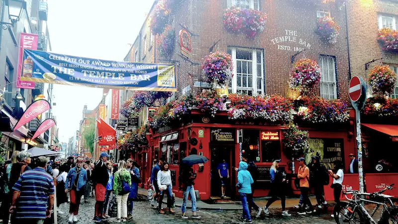 People walking around in rain jackets with a brightly decorated Temple Bar behind at the start of the street of bars in Dublin, Ireland.