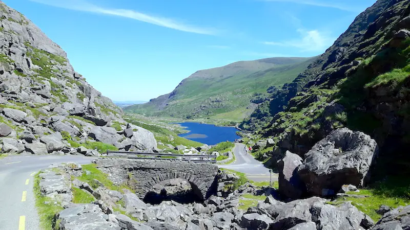 Winding road over a stone bridge with a blue lake in the middle of green hills in Gap of Dunloe, Ireland.