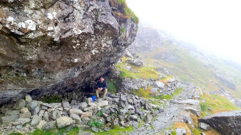 Man taking shelter under a huge boulder on a wet, cloudy hiking trail in Llyn Ogwen, Wales.