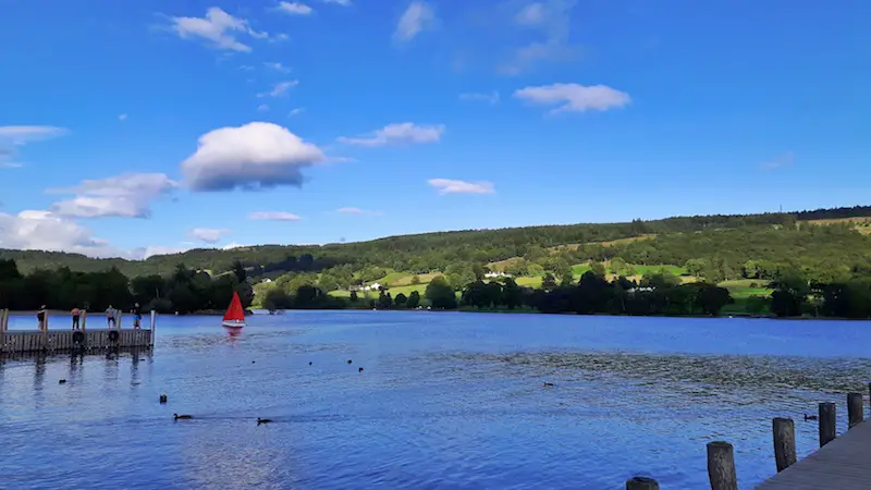 Red sailboat on a blue lake surrounded by green hills in Coniston Waters, Lake District, England.