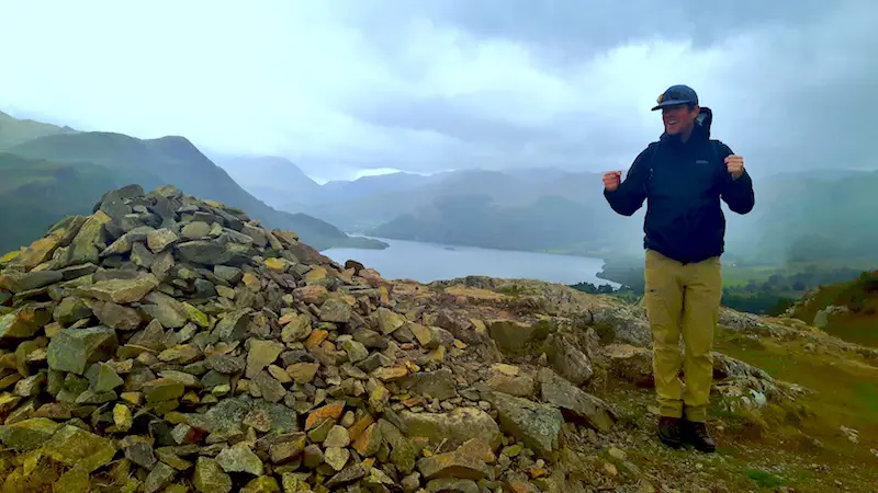 Man celebrating reaching Yew Crag summit in Ullswater, Lake District with view to the lake on a stormy day in England.