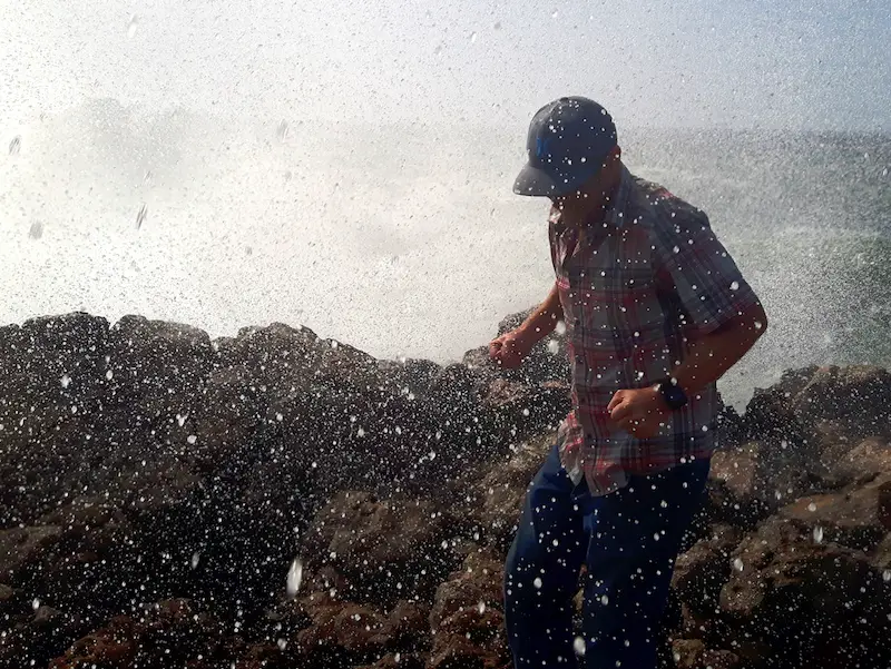 Man getting soaked from a surprise wave crashing over sharp rocks in Bordeira, Alentejo region, Portugal.