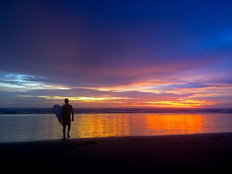 Brilliant oranges, purples and blues at sunset with the silhouette of a man carrying a surfboard out of the water in Nosara, Costa Rica.