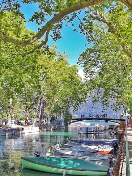 Row of canal boats with a bridge over entrance to Lake Annecy, surrounded by French Alps.