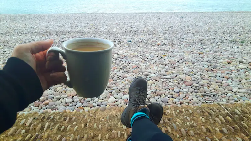 Hiking boots and a coffee on a pebble beach in Budleigh Salterton, Devon England.