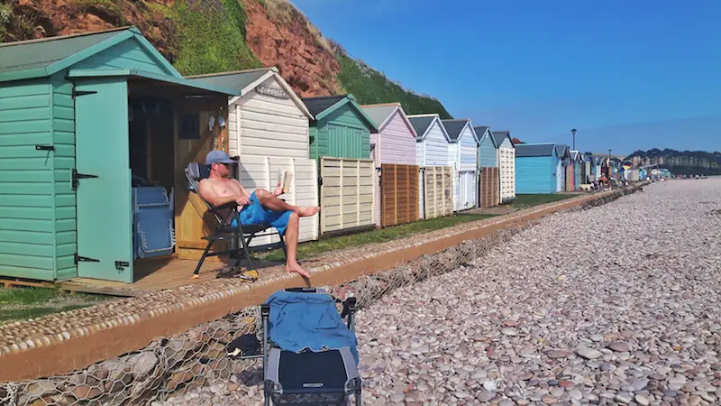 Man reading outside an open beach hut in a row of beach huts on the pebble beach in Budleigh Salterton, Devon England.