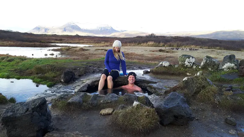 Man in a hole in the ground with a woman sitting on the edge of a hot pot in a field in Iceland.