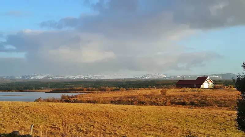 Brown grass fields lead to a lake surrounded by mountains with one lone red and white building on the shore in Iceland.