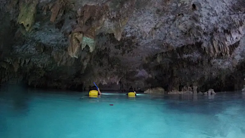 Two people in life jackets swimming into the entrance to a cenote near Tulum