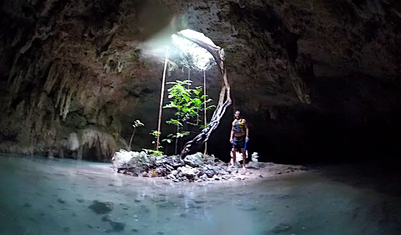 Inside a cave with a hole in the roof and tree roots coming in. Turquoise water all around in Cenote Sac Actun