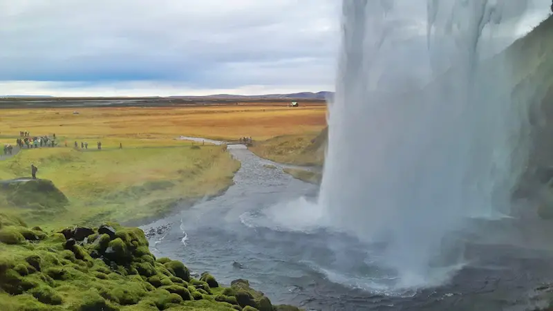 looking from behind a huge waterfall with green moss, and fall coloured fields, Seljalandsfoss, Iceland