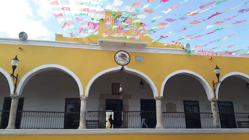 Yellow building with large arches and brightly coloured banners strung across the street in Izamal, Yucatan Mexico.