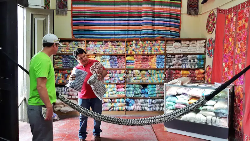 A Mayan artisan showing a man a selection of hammocks with a wall filled with bagged hammocks behind him in Merida, Mexico.