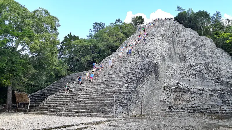 Tall stone pyramid surrounded by trees with people climbing to the top at Coba ruins, Mexico.